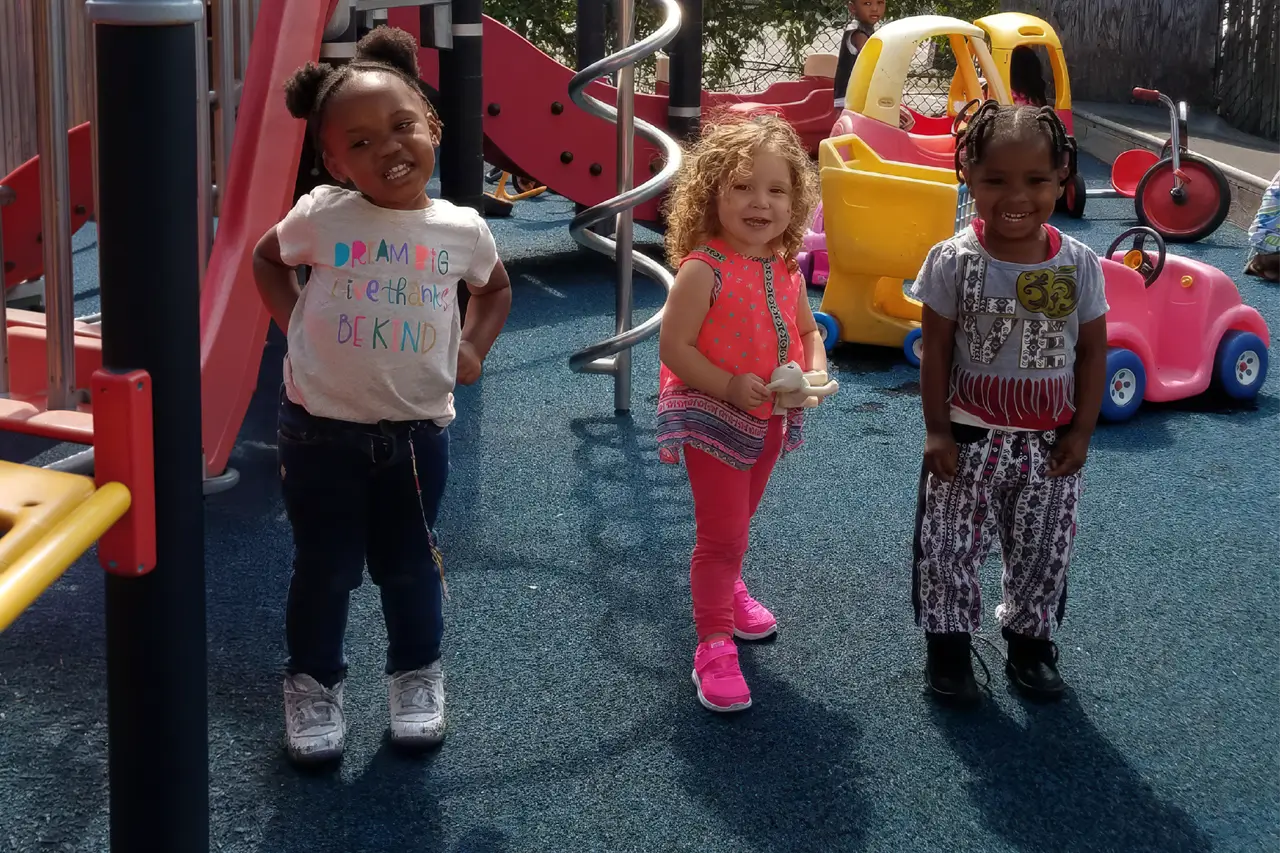 Three children smile while standing in front of colorful playground equipment, ready to play.